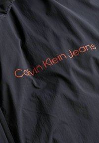Juodas švarkas, pagamintas iš lengvos medžiagos, su lygiu paviršiumi. Išryškinta „Calvin Klein Jeans“ logotipas oranžine spalva.