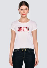 Žena se rovnými vlasy, nosící zlaté kruhové náušnice, bílý tričko s nápisem "BIG STAR", černým páskem a tmavě modrými džíny, stojí na jednobarevném pozadí.