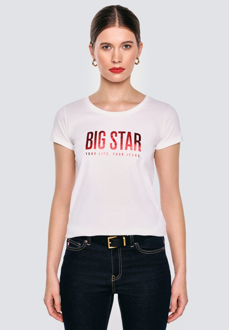 Žena se rovnými vlasy, nosící zlaté kruhové náušnice, bílý tričko s nápisem "BIG STAR", černým páskem a tmavě modrými džíny, stojí na jednobarevném pozadí.