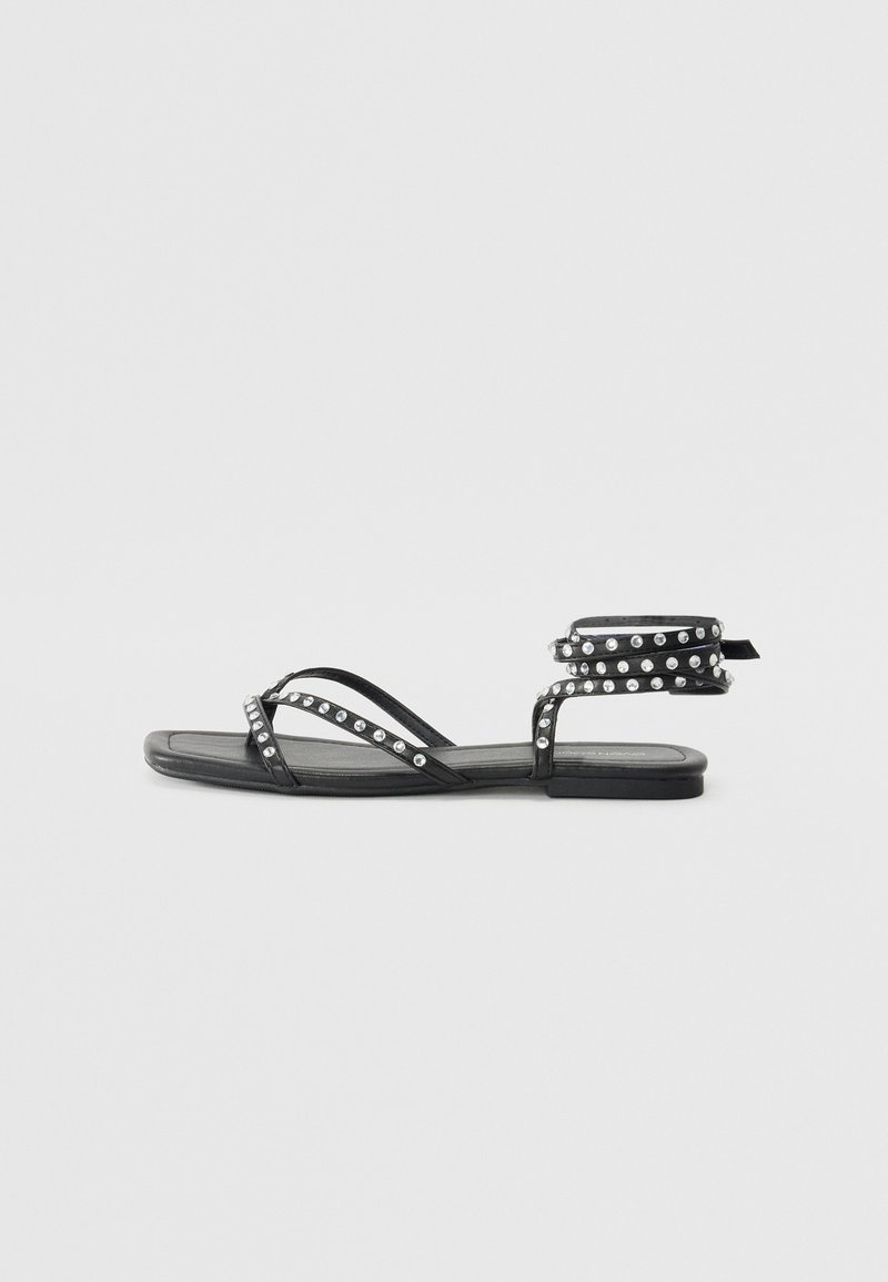 Sandalele plate negre au curele subțiri împodobite cu nituri argintii, un design minimalist și o curea pentru gleznă cu un mecanism de prindere.
