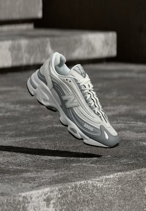 Scarpa da ginnastica New Balance 1000 bianca e grigia che fluttua sopra una superficie in cemento con scale sullo sfondo.