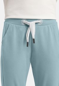Helle blaue Jogginghose mit einem Kordelzugbund, ausgestattet mit weißen Details und tonalen Nähten. Enthält zwei Seitentaschen. Glatte Textur.