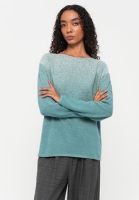 Texturiertes Langarm-Sweatshirt in einem Farbverlauf von Teal, mit einem Boot-Ausschnitt und gerippten Bündchen, kombiniert mit grauen karierten Hosen.
