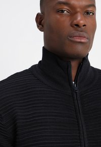 Svart zip-up högkragad tröja med texturerat ribbat material, kroppsnära design och slät dragkedjedetalj.