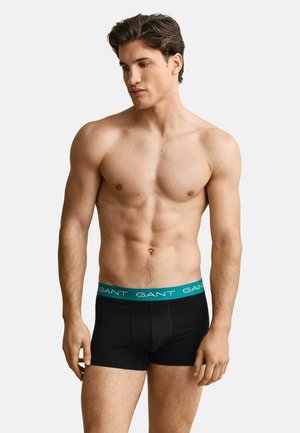 Mann mit kurzen dunklen Haaren, der schwarze Boxer-Shorts mit einem türkisfarbenen Bund trägt, auf dem der Markenname "GANT" steht, und vor einem schlichten Hintergrund steht.