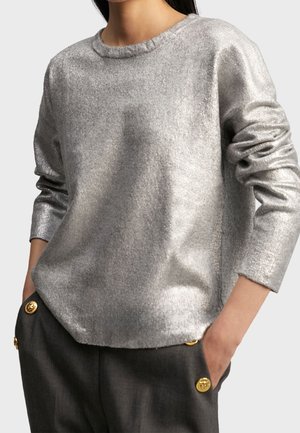 Vrouw draagt een metallic zilverkleurige lange mouw trui, ingestopt in donkergrijze broek met gouden knoopdetails, handen in de zakken.