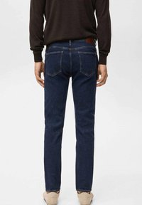 Person trägt dunkelblaue Slim-Fit-Jeans und ein schwarzes Langarmshirt, steht mit entspannten Armen an den Seiten vor einem weißen Hintergrund.