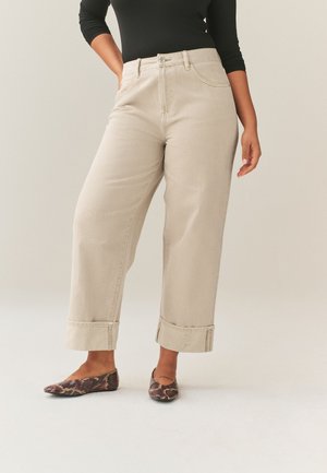 Žena oblečená v béžových širokých krátkych nohaviciach s ohrnutými lemami a plochých topánkach so vzorom hadieho vzoru, stojaca pred jednoduchým pozadím.