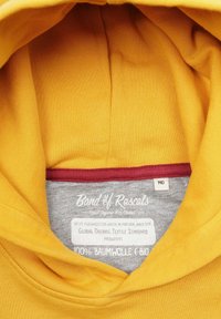 Gul hoodie-interiör med en grå etikett med text. Materialet är 100 % ekologisk bomull. Röd detalj vid huvens kant. Storleksmärkning visar 140.