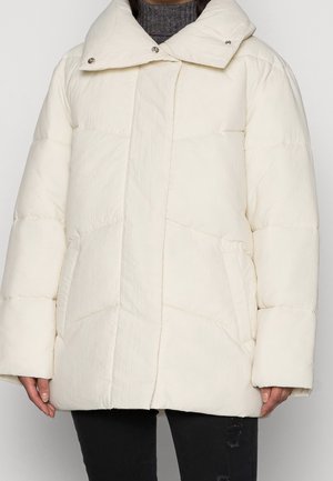 Vrouw die een off-white pufferjack met hoge kraag draagt over een grijze coltrui en zwarte gescheurde spijkerbroek, staand tegen een effen achtergrond.