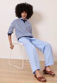 Blå- och vitrandig tröja med V-ringning, parat med ljusblå randiga byxor. Bruna sandaler och en trådstol i en minimalisk miljö.