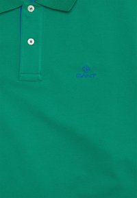 Polo en coton vert avec un col classique, une patte de boutonnage à trois boutons et un logo "GANT" brodé en bleu sur la poitrine gauche.