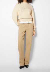 Beige neulepaita, jossa on teksturoitu kuvio ja pitkät hihat, yhdistettynä beigeihin housuihin ja mustiin sileäpintaisiin kengiksi, joissa on terävä kärki.