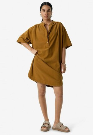 Robe à manches courtes de couleur moutarde avec un col à patte et un ourlet arrondi. Portée avec des sandales texturées, mettant en valeur un ajustement confortable.