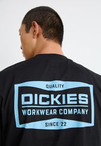 Svart bomull t-skjorte med en stor blå rektangulær logo på baksiden med teksten "DICKIES WORKWEAR COMPANY" og "KVALITET Siden '22."