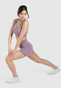 Fioletowy sportowy crop top i szorty z wysokim stanem, wykonane z rozciągliwego materiału o gładkiej fakturze. Model w pozycji wykroku, noszący białe sneakersy.