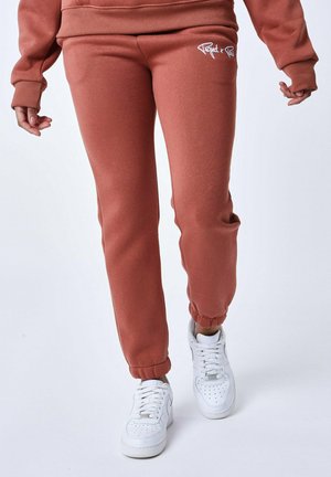 Pantalons joggers couleur rouille en tissu doux, dotés de poignets élastiques, d'un petit logo blanc à l'avant, et associés à des baskets blanches.