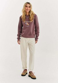 Bordowa bluza z nadrukiem, kremowe spodnie oraz brązowe buty trekkingowe z różowymi sznurówkami. Swobodny strój odpowiedni na outdoor.