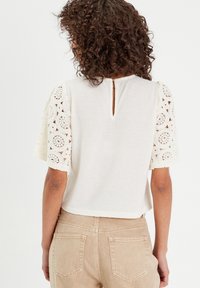 Haut ivoire à manches courtes avec des manches en dentelle, détail en forme de clé ronde dans le dos et une coupe décontractée. Assorti avec un pantalon beige clair.