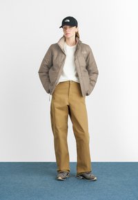 Tan insulated jacket, white sweatshirt, beige pants, black cap. The jacket has a smooth texture, and pants feature a relaxed fit and button closure.
