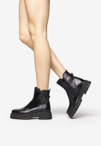 Black leather ankle boots with a textured back, buckle detail, and a thick, rugged sole. Smooth finish and simple, elegant design.