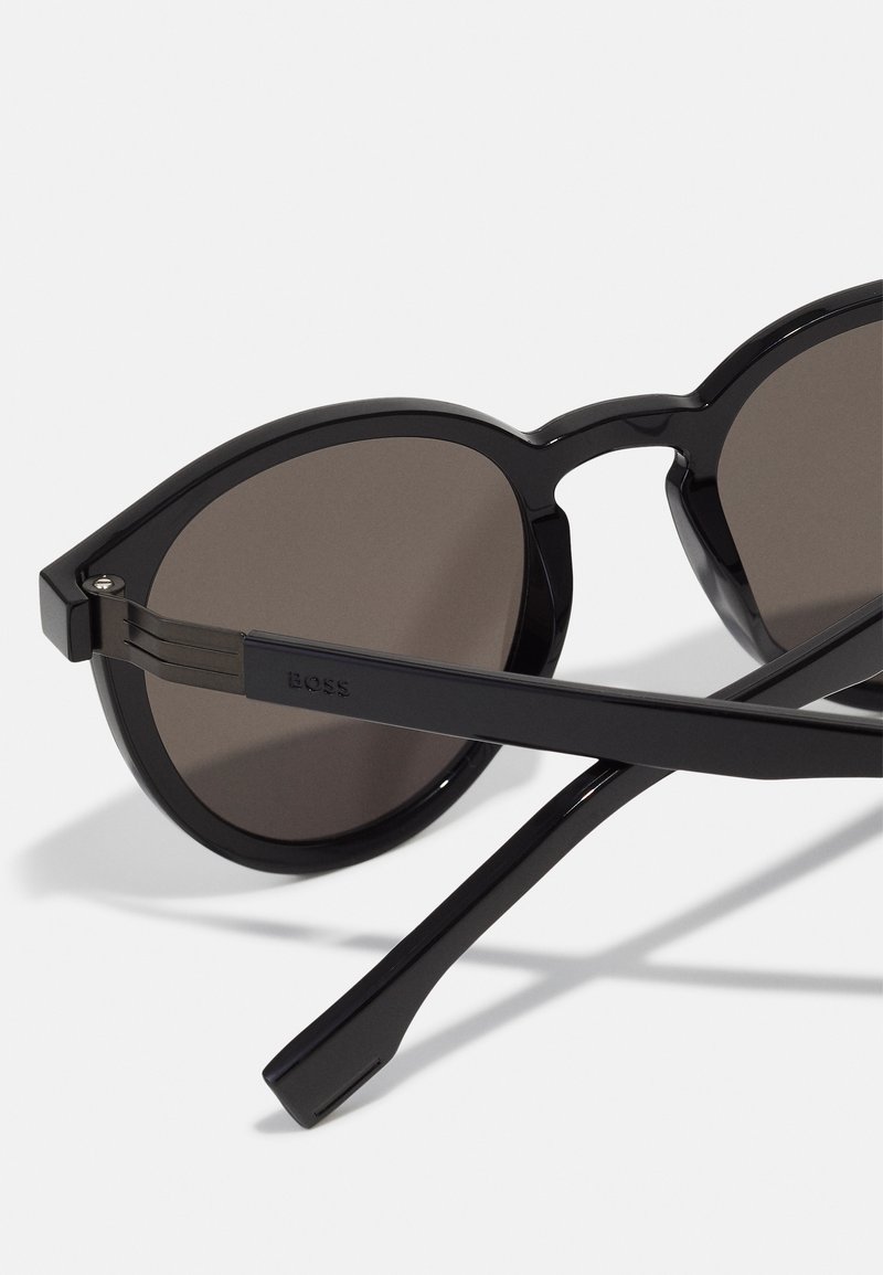 Occhiali da sole neri con montatura arrotondata, lenti grigie e astine sottili e angolari. Presentano il marchio "BOSS" all'interno dell'asta.