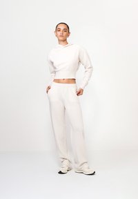 Persoană stând cu mâinile în buzunare, purtând un hanorac scurt complet alb, pantaloni largi și adidași albi, pe un fundal simplu.
