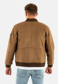 Giacca bomber in suede marrone con colletto a coste, caratterizzata da una texture liscia e una vestibilità casual. Include un patch con il marchio sulla manica.