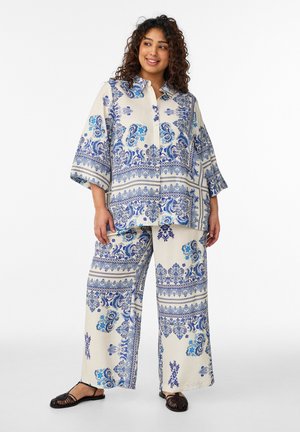 Kvinde stående iført løstsiddende cremefarvet tøj med blå paisley- og blomsterprint, kombineret med sorte flade sandaler i vævet materiale.