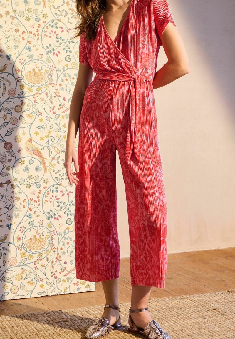 Femme portant une combinaison-pantalon cache-cœur rouge à manches courtes et motifs, nouée à la taille, debout sur un tapis avec un papier peint fleuri en arrière-plan.