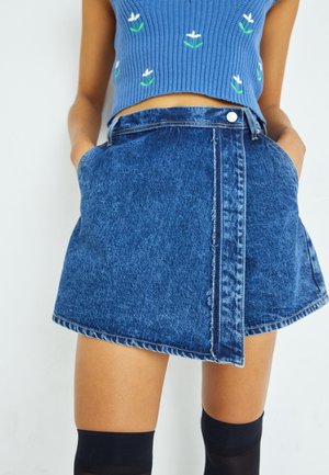 Niebieski dzianinowy krótki top w białe kwiaty, zestawiony z niebieską, mini spódnicą z denimu o wysokim stanie oraz czarnymi pończochami sięgającymi ponad kolana, na stojącej osobie.