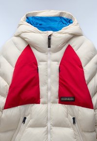 Giacca puffer color crema con audaci accenti triangolari rossi. Presenta un rivestimento interno della cappuccio blu e tasche con zip. Design testurizzato e isolato.