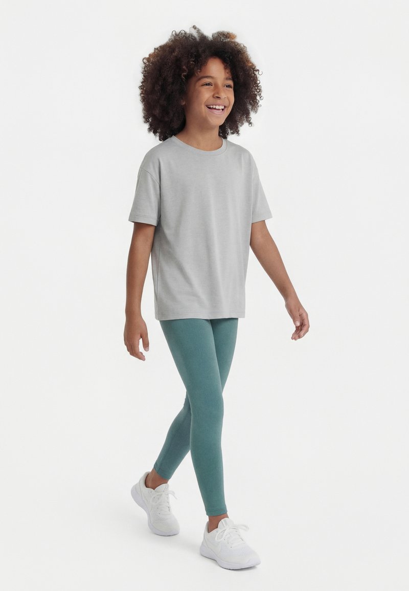 Χαμογελαστό παιδί με σγουρά μαλλιά που περπατά, φοράει φαρδιά γκρι μπλούζα, τιρκουάζ κολάν και λευκά αθλητικά παπούτσια σε απλό λευκό φόντο.