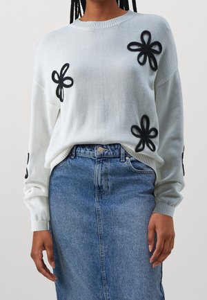 Witte trui met zwarte bloemapplicaties, geribbelde hals en mouwen, gecombineerd met een blauwe denimrok. Zachte textuur, losse pasvorm.