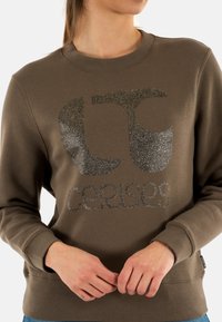Felpa marrone con scollatura rotonda, caratterizzata da un design del logo glitterato in varie sfumature di grigio e un orlo regolabile. Tessuto morbido.