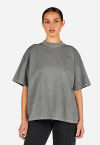 Hallhalli ülikerge hall T-särk pehme puuvillaga, kõrge kaelusega, vajunud õlgadega ning diskreetse tikandiga logo rinnal.