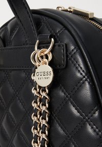 Sac à main en simili cuir noir matelassé avec une bandoulière en chaîne dorée et un pendentif rond portant l'inscription "GUESS EST.1981" en relief. Détails supplémentaires : fermeture éclair.