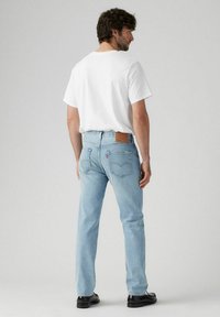 Jeans de mezclilla azul claro con corte recto y un parche de cuero en la parte trasera, combinados con una camiseta blanca sencilla, vistos desde la parte de atrás. Zapatos negros.