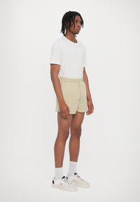 Beige Shorts mit elastischem Bund und ein eng anliegendes weißes T-Shirt mit kurzen Ärmeln, kombiniert mit weißen Sneakern und grauen Socken. Einfache und funktionale Gestaltung.