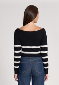Maglione a righe bianche e nere con design off-shoulder. Caratterizzato da maniche lunghe e una vestibilità cropped, abbinato a jeans blu a vita alta.