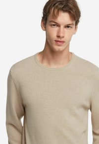 Beige stickad tröja med långa ärmar och rund halsringning. Mjuk textur och subtil räfflad mönster på manschetter och fåll.