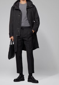 Schwarzer langer Mantel mit Kapuze über einem grauen T-Shirt, kombiniert mit maßgeschneiderten schwarzen Hosen und schwarzen Lederschuhen. Schwarze Handtasche in der Hand.