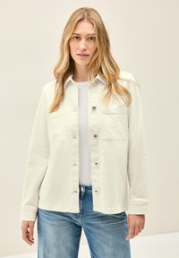 Valkoinen kevyt takki, jossa on kaksi rintataskua, hopeiset napit ja hieman teksturoitu pinta, päällä valkoinen paita ja siniset farkut.