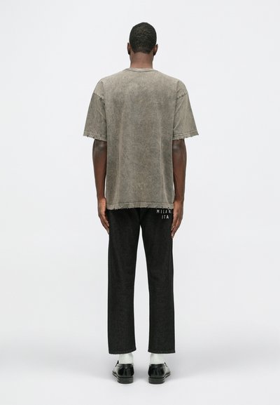 Tricou oversized din in gri cu margini destrămate, purtat împreună cu pantaloni din denim negri. Pantalonii au inscripționat cu alb textul "MILANO ITA".
