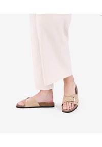 Sandali slide beige chiaro realizzati in materiale morbido simile alla suede, con una fascia ampia e una fibbia argentata, e una suola in sughero texturizzata.