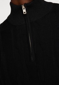 Black knit sweater with a high collar, featuring a half-zip closure, textured cable pattern, and ribbed cuffs.