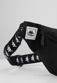 Czarna nerka wykonana z trwałego materiału, z wyraźnym logo Kappa, regulowanym paskiem z białymi logo oraz zamknięciem na zamek.