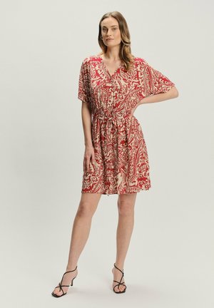 Kobieta w krótkiej sukience w czerwono-beżowy wzór paisley z wiązaniem w talii oraz czarnych sandałach na obcasie, stoi z jedną ręką na biodrze.