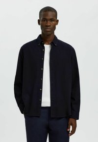 Granatowa koszula z długim rękawem, wykonana z miękkiego materiału, z klasycznym kołnierzykiem i złotymi guzikami, nałożona na białą koszulkę.