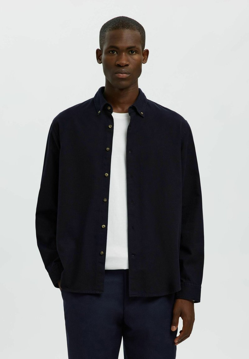 Granatowa koszula z długim rękawem, wykonana z miękkiego materiału, z klasycznym kołnierzykiem i złotymi guzikami, nałożona na białą koszulkę.